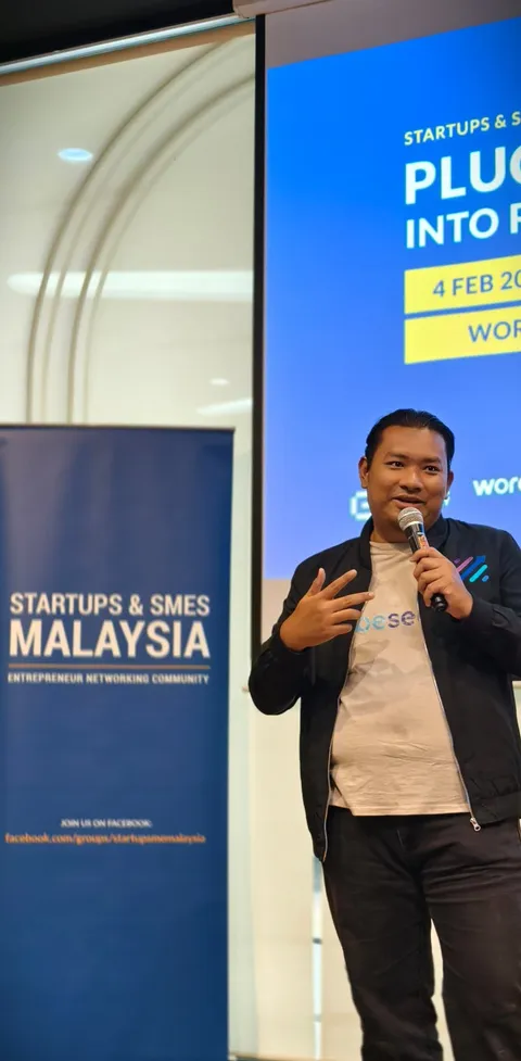 Hafiz Zainudin presenting at Growth Charger event, speaking into microphone with business metrics presentation in background
